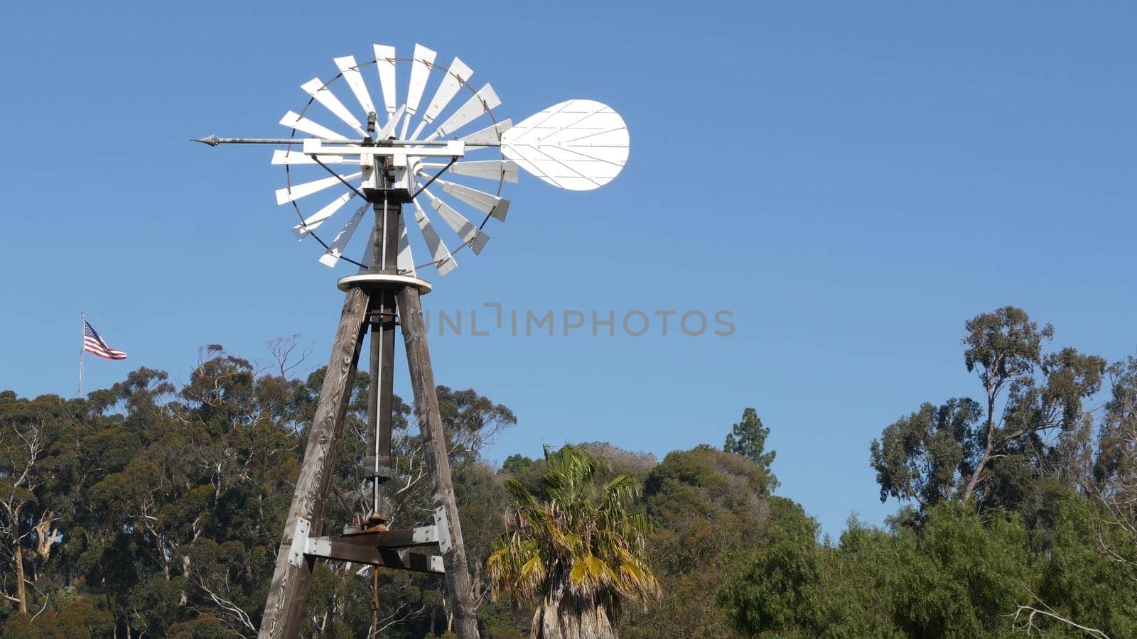 Classic retro windmill, bladed rotor and USA flag against blue sky. Vintage water pump wind turbine, power generator on livestock ranch or agricultural farm. Rural symbol of wild west, rustic suburb.