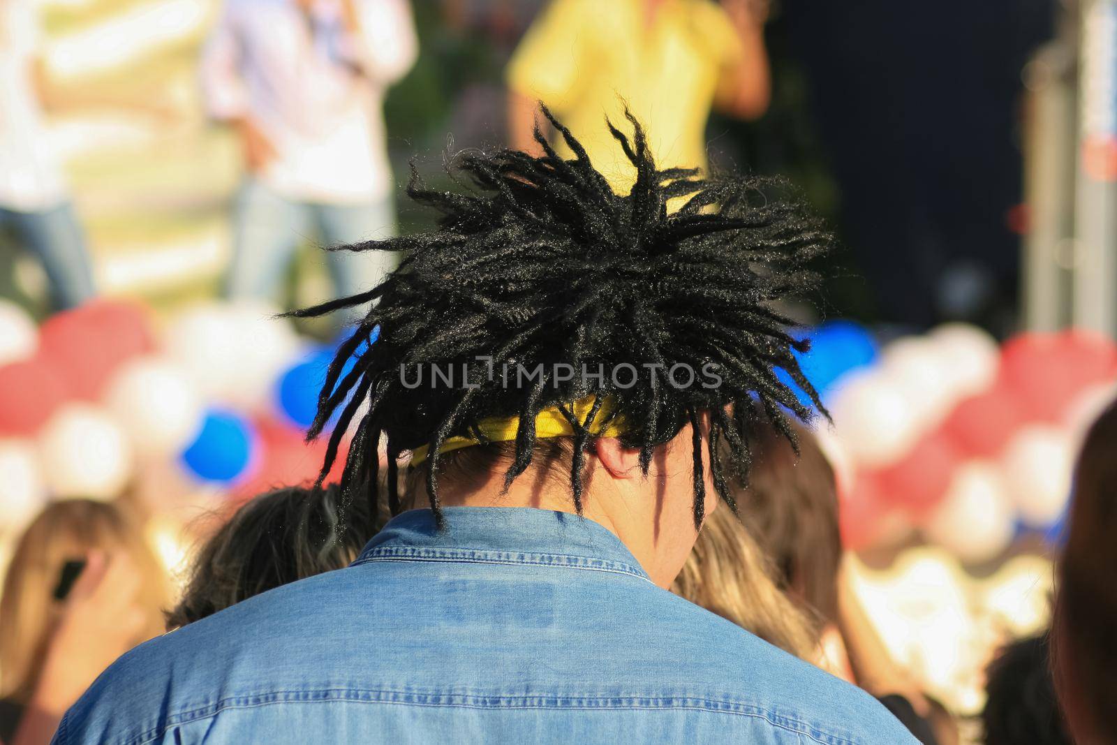 A man in a black dreadlock wig at a street concert. Stands with his back, looks to the side. Sun, summer.