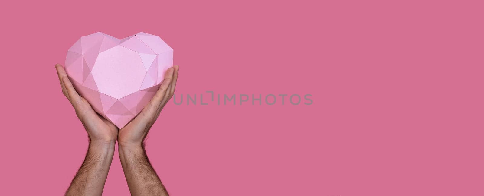 Polygonal three-dimensional pink heart made of paper in the hands of a man. On a pink background. Banner. Copy space. 