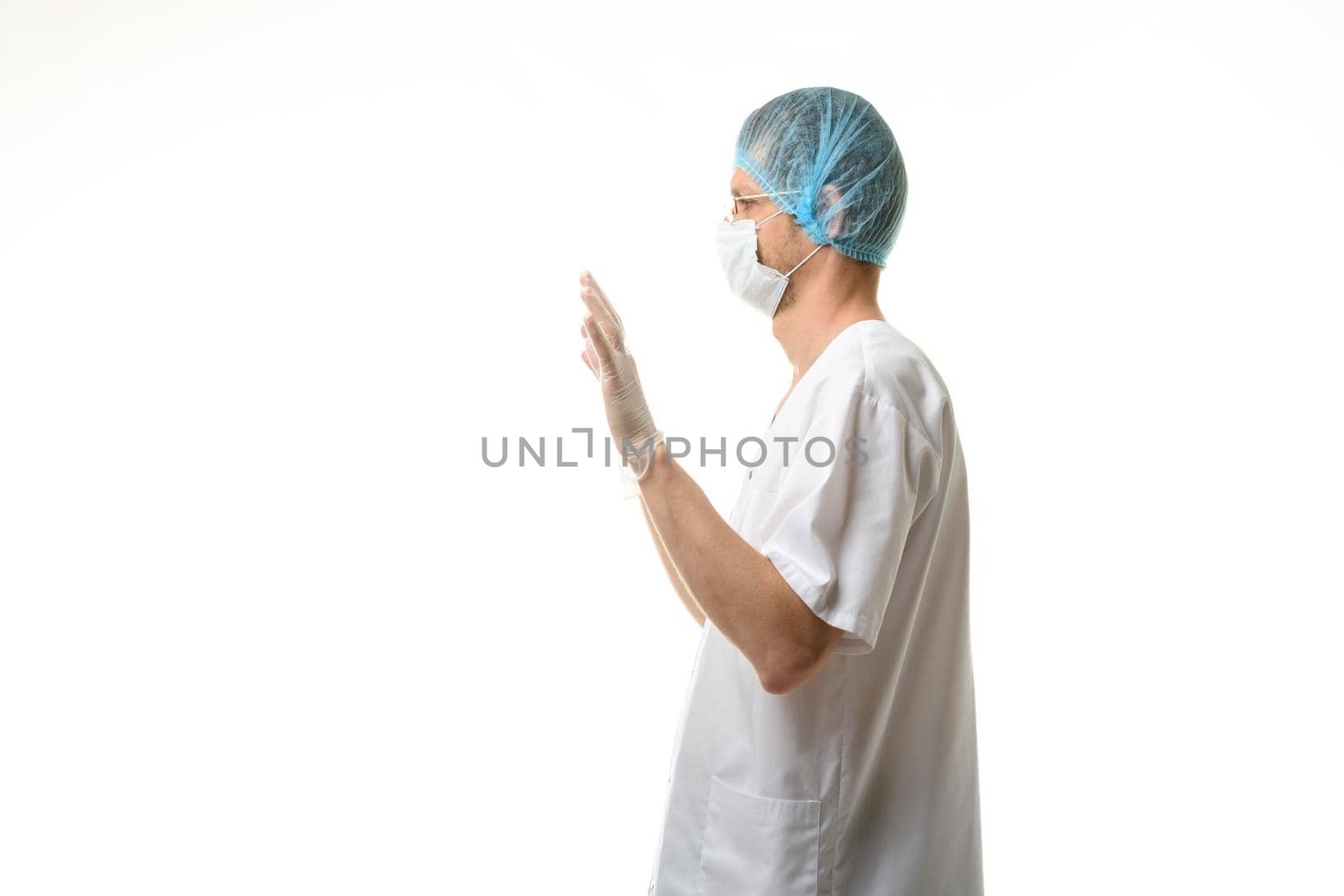 A male doctor stands with his hands raised up, gloves are on, a mask and a hat are on his head, side view