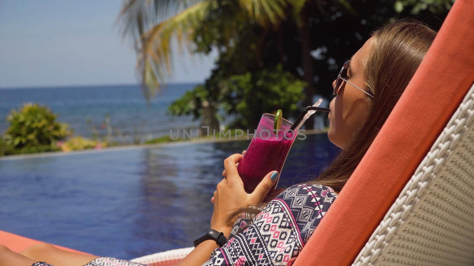Beautiful young girl with glass of cocktail relaxing near swimming pool. Girl in sunglasses with cocktail in seafront near the bassin amongst the palm trees on the background of the sea. Travel, vacation concept.