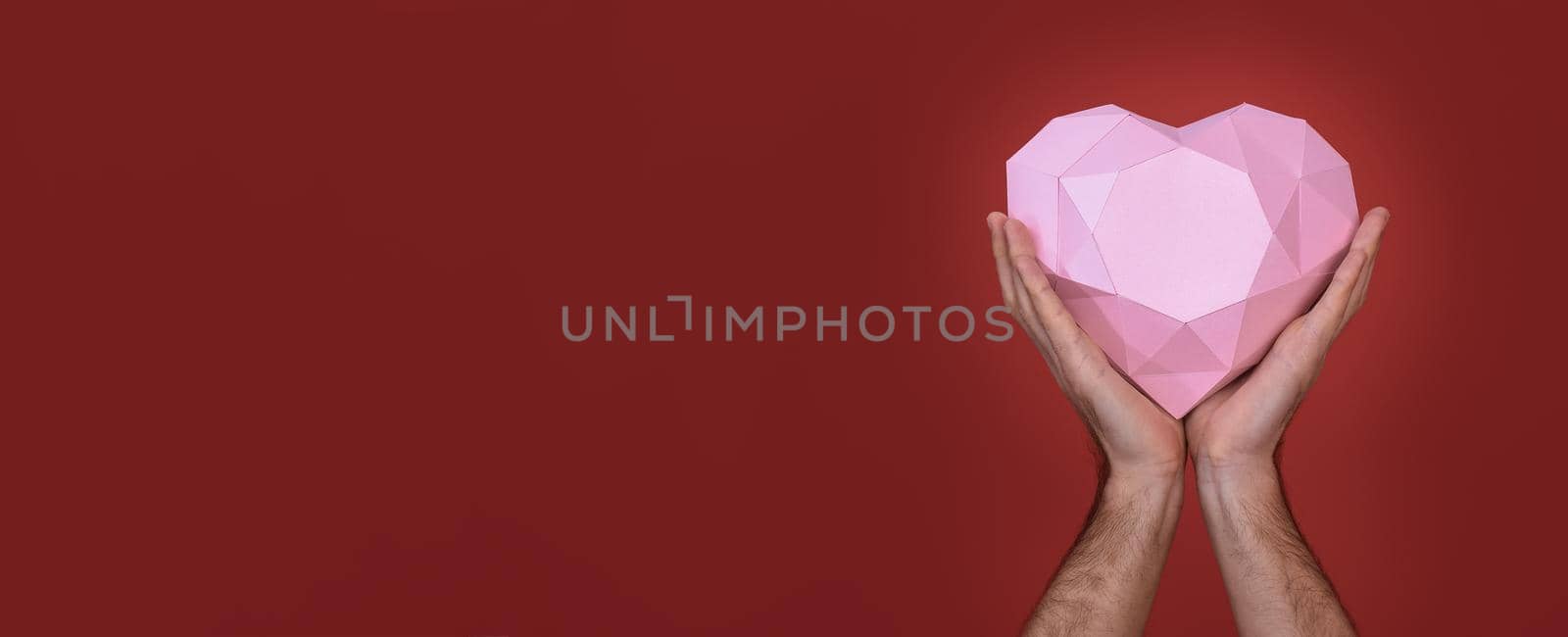 Polygonal pink heart in the hands of a man close-up. A banner with space to copy. On a red background. Greeting cards for Valentine's Day, weddings, declarations of love. 