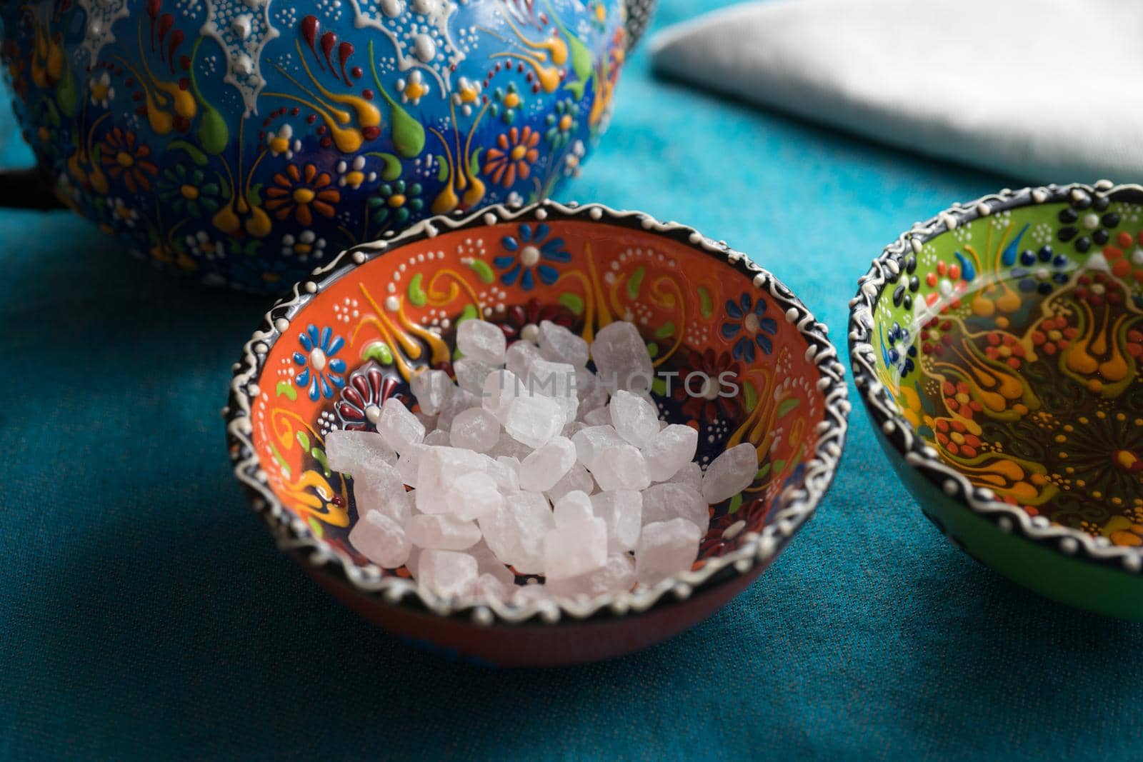 Large crystals of white sugar in colorful patterned ceramic bowl in turquoise textile tablecloths.