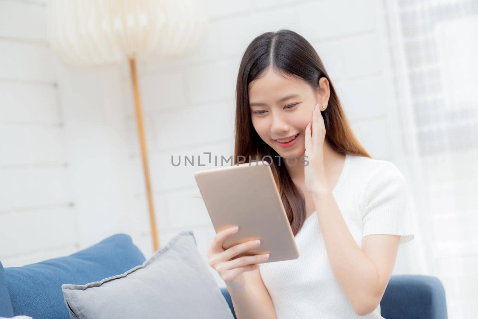 Young attractive asian woman resting using browsing tablet computer on sofa at home, happy girl sitting on couch relax reading digital gadget at house, communication and lifestyle concept.