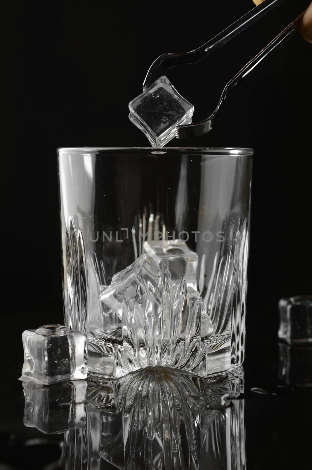 A hand adding some fresh ice with tongs into a clean whiskey glass over a black reflective background.