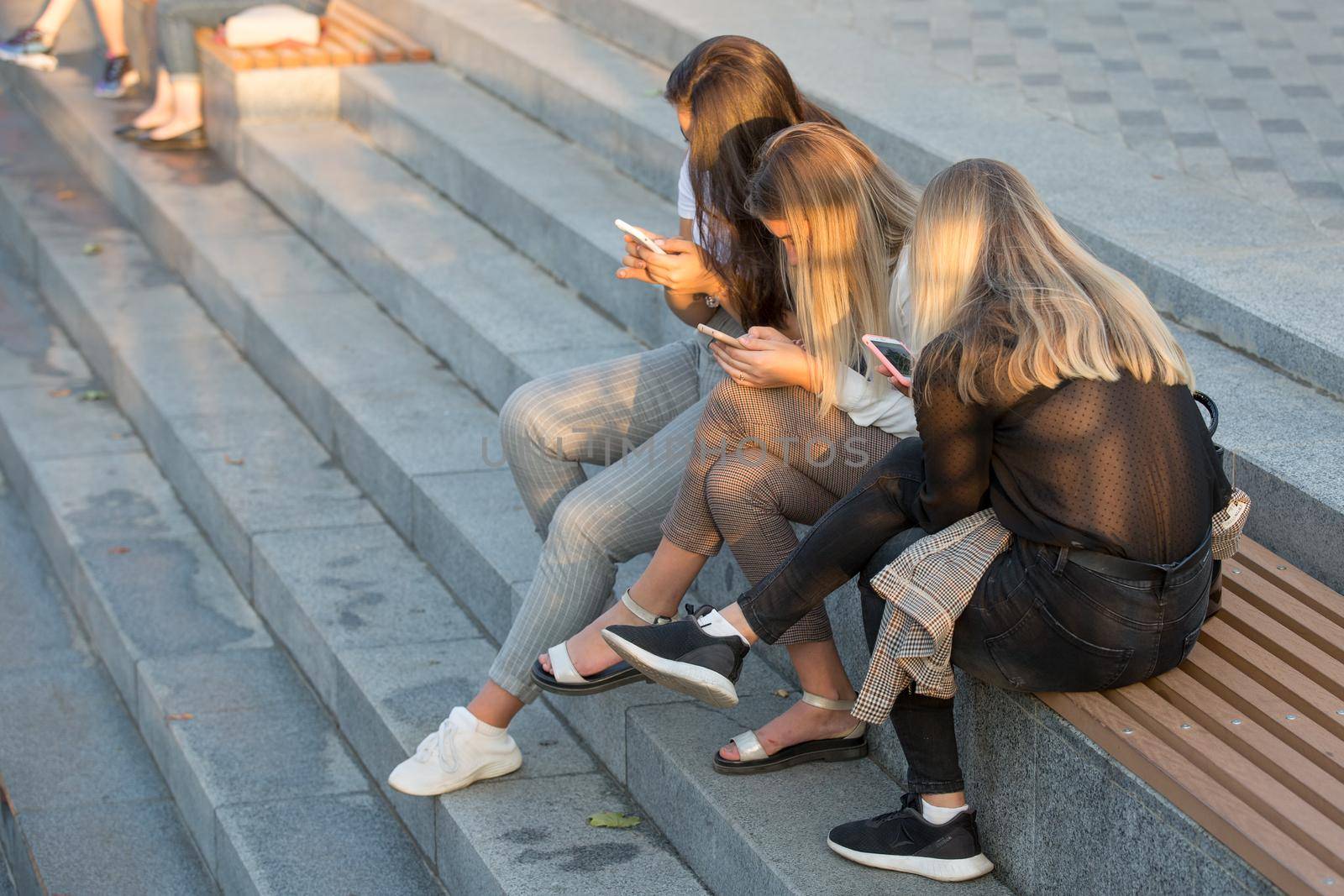 Youth browsing social networks. Girls are holding mobile phones. Three girls in a city park use gadgets. Modern generation and telecommunications.