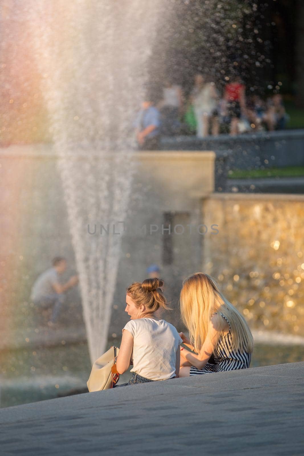 Young people are sitting in the park. Youth peacefully resting in city parks. Hot summer evening. Rainbow over the fountain. Weekend in the city park. Healthy rest.