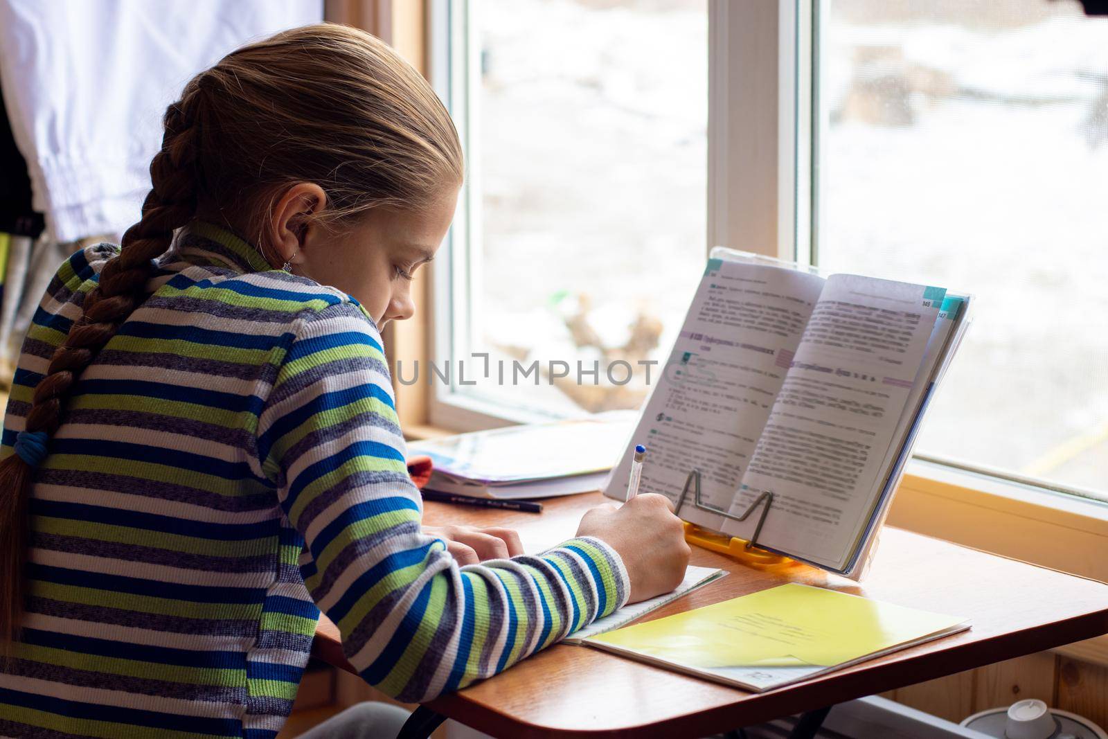 Schoolgirl sits at the table by the window and does homework