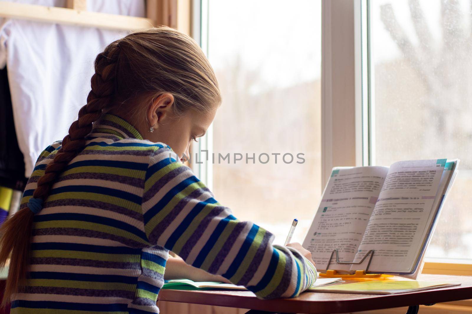 Schoolgirl sits at a table by the window and does her homework, view from the back