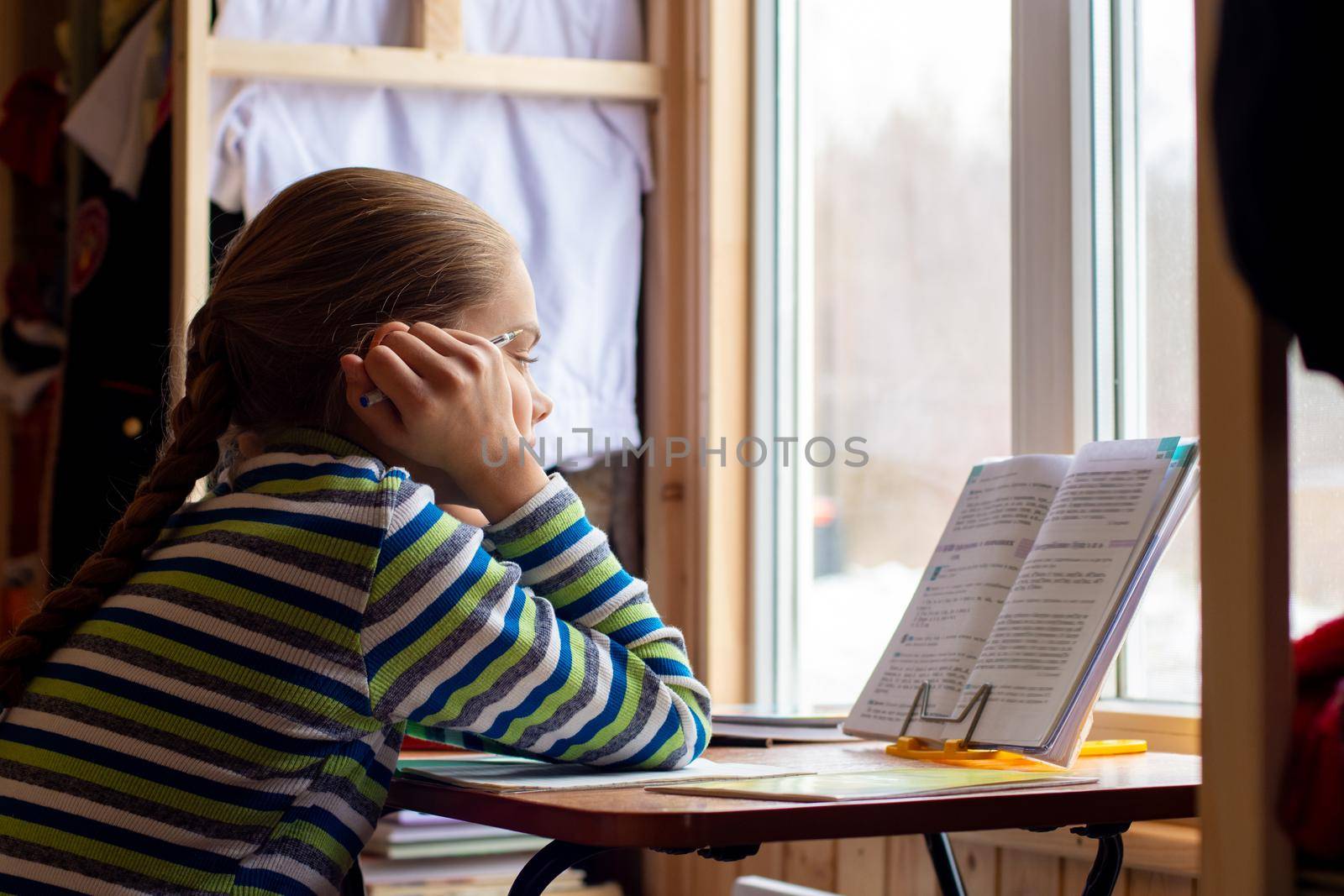 The schoolgirl carefully reads the task in the textbook while sitting at the table by the window at home