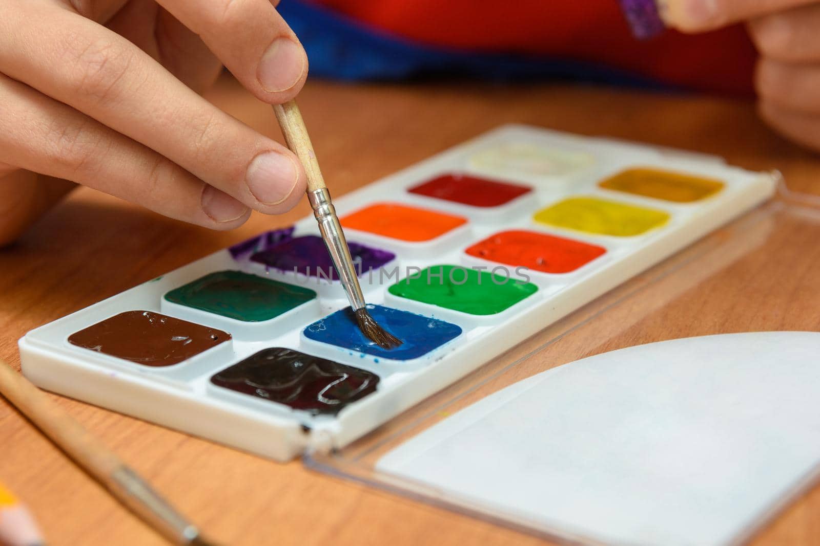 The child's hands are dipping with a brush in the desired color of watercolor paints