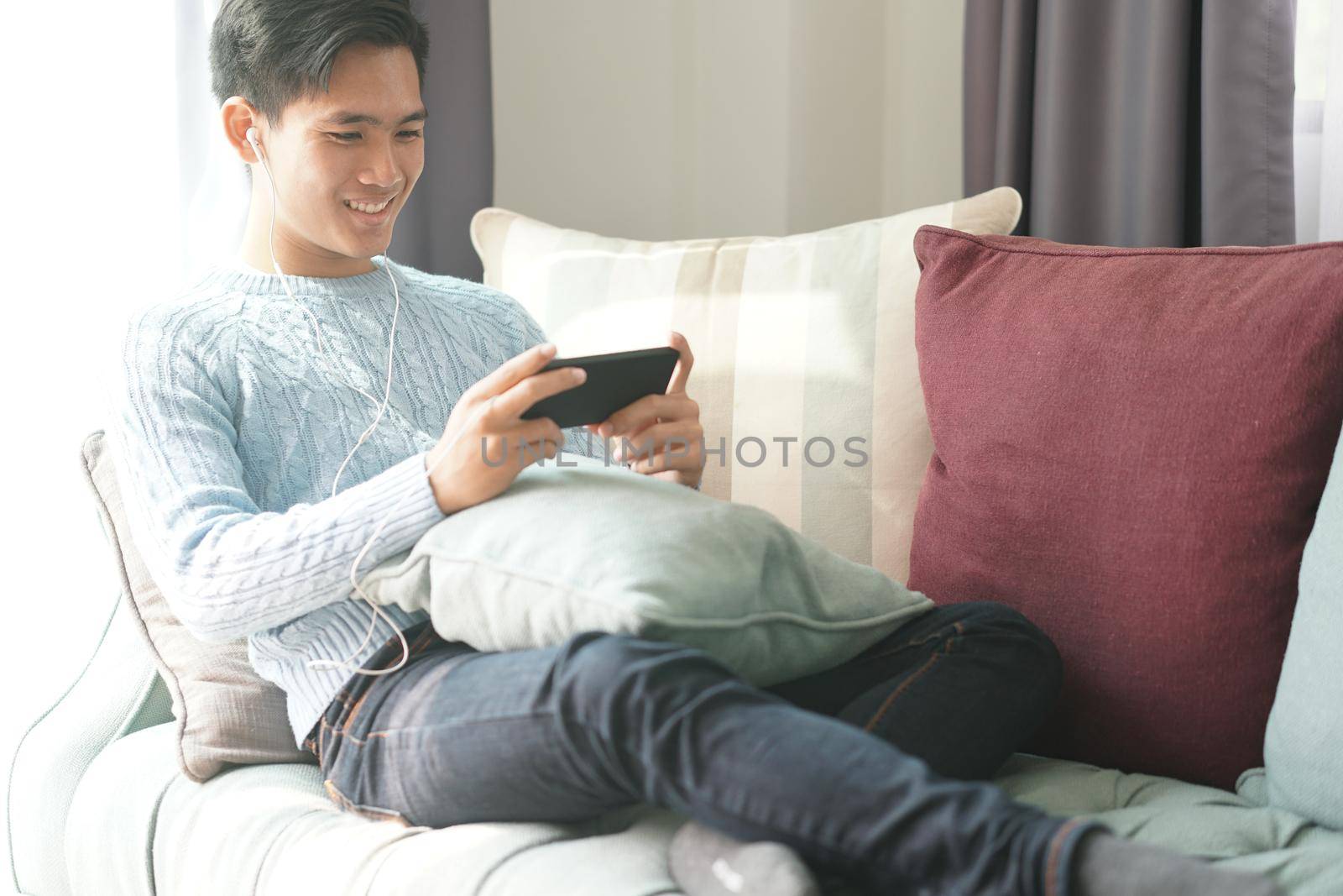 young asian man wearing earphones listening to music on smart phone relaxing on sofa at home