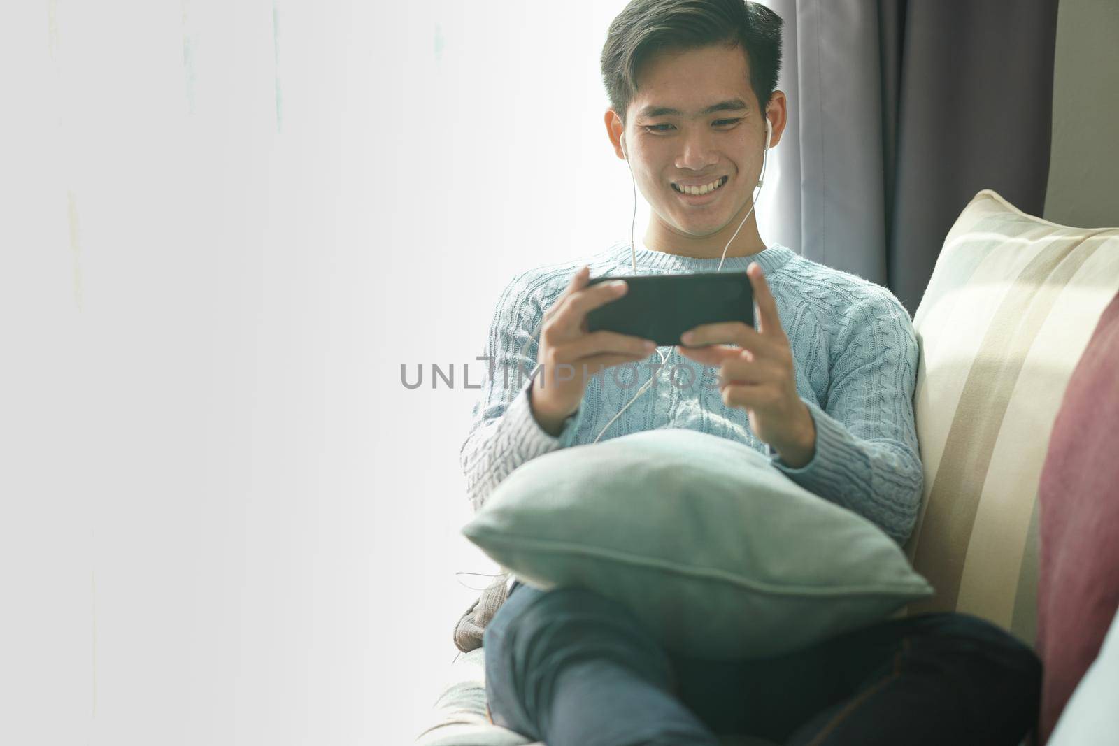 young asian man wearing earphones listening to music on smart phone relaxing on sofa at home