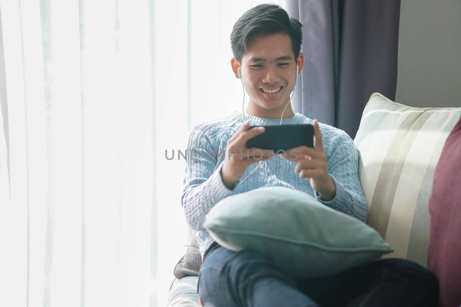 young asian man wearing earphones listening to music on smart phone relaxing on sofa at home