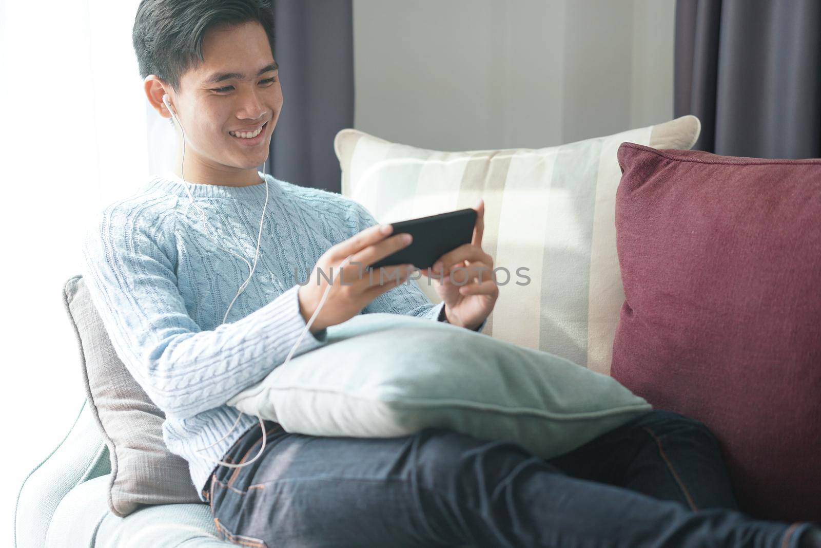 young asian man wearing earphones listening to music on smart phone relaxing on sofa at home