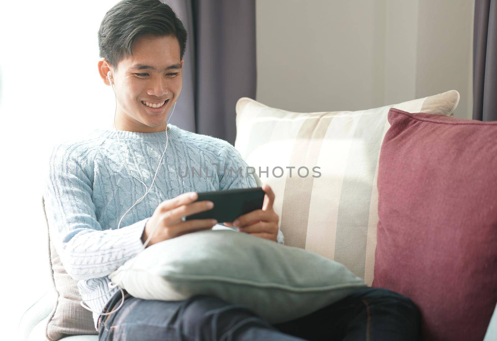 young asian man wearing earphones listening to music on smart phone relaxing on sofa at home