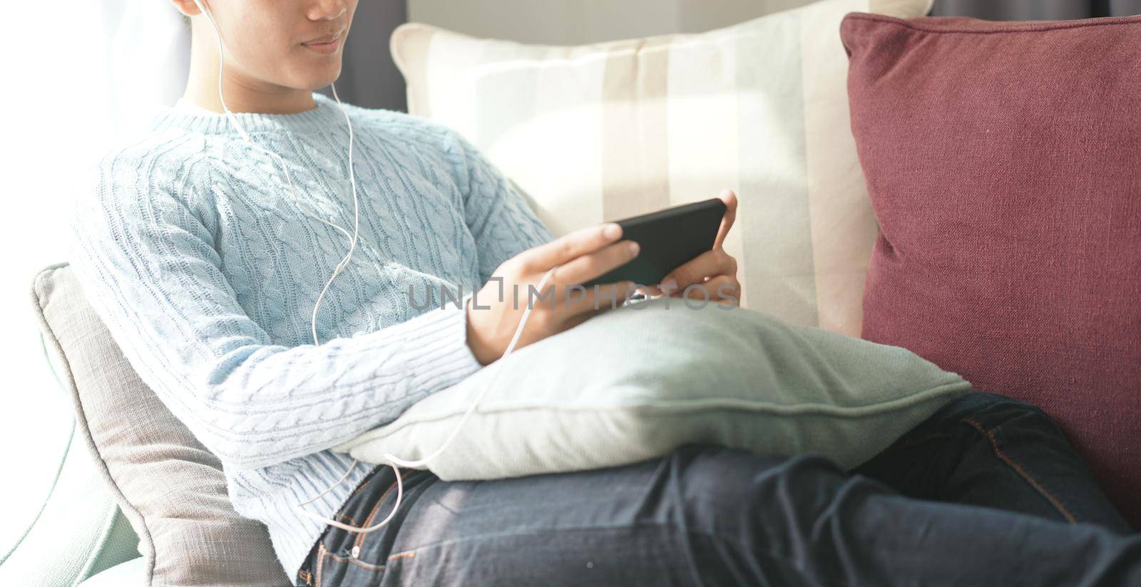 young asian man wearing earphones listening to music on smart phone relaxing on sofa at home