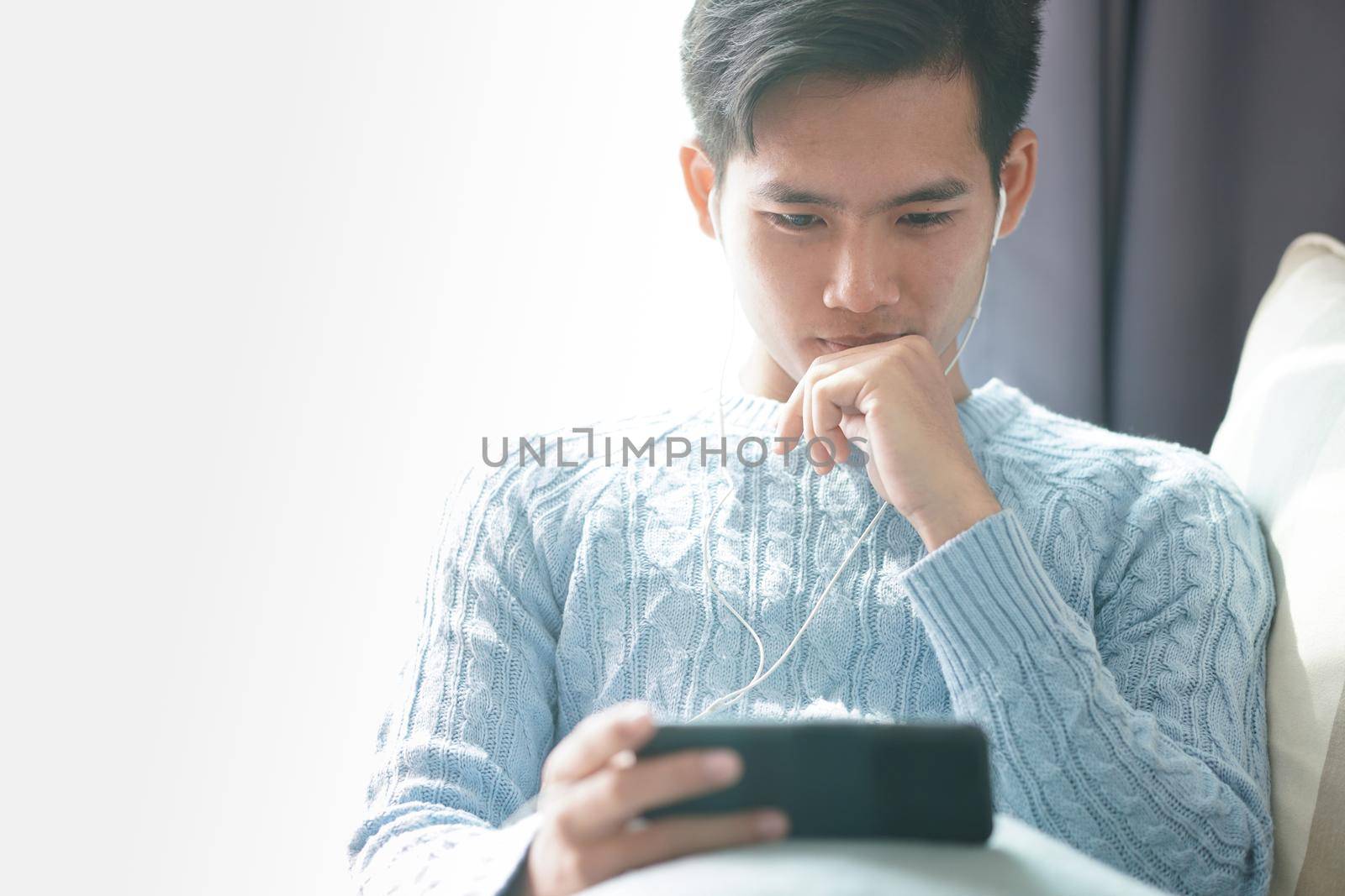 young asian man wearing earphones listening to music on smart phone relaxing on sofa at home