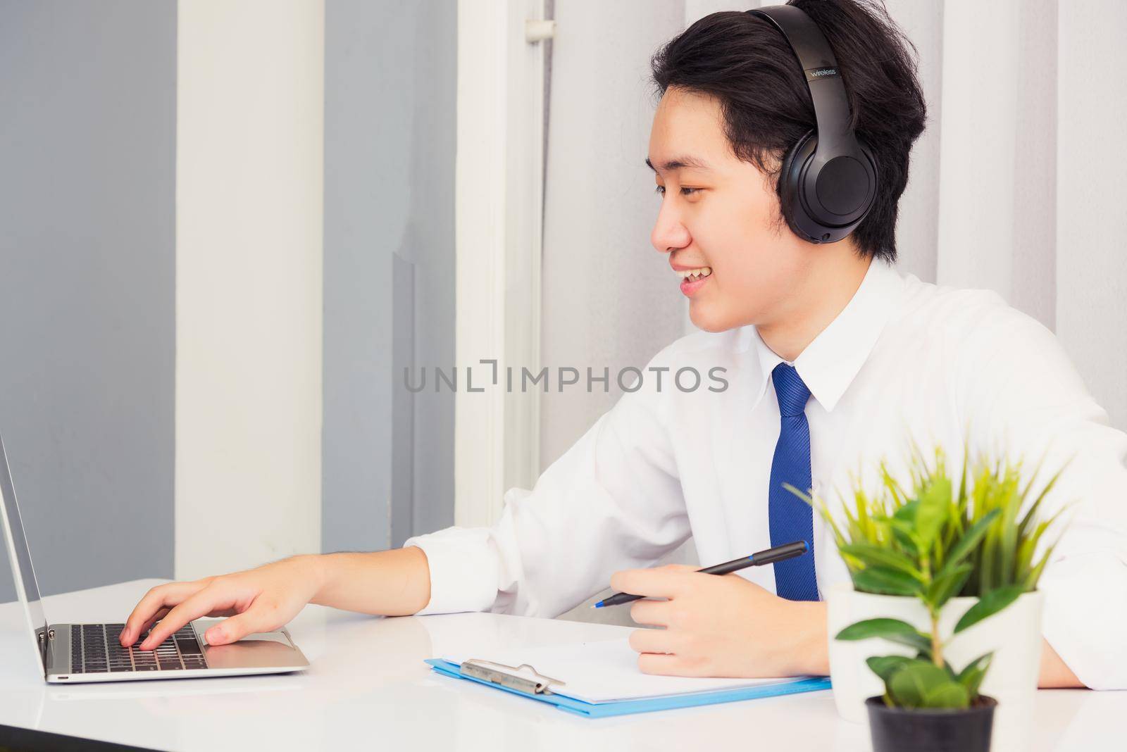 Work from home, Asian young businessman wear suit video conference call or facetime he smiling sitting on desk using laptop computer and write paper notebook information at home office