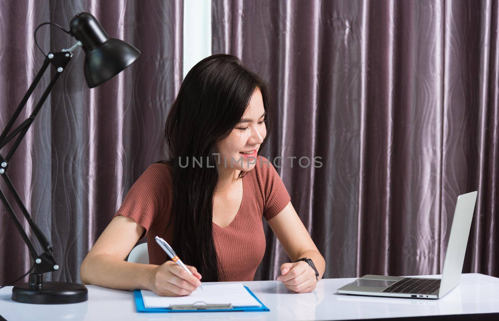 Work from home, Asian young businesswoman smile video conference call or facetime by laptop computer meeting with team and her writing on paper noted on desk at home office