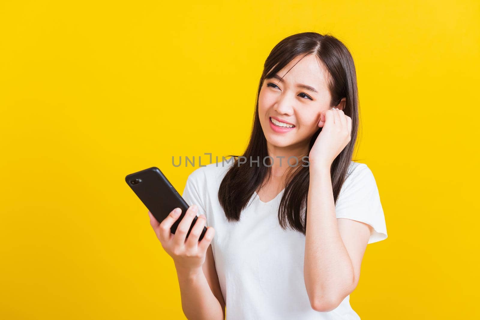 Portrait Asian of a happy beautiful young woman holding a mobile smart phone and wearing wireless headphones listening to music from smartphone studio shot isolated on yellow background