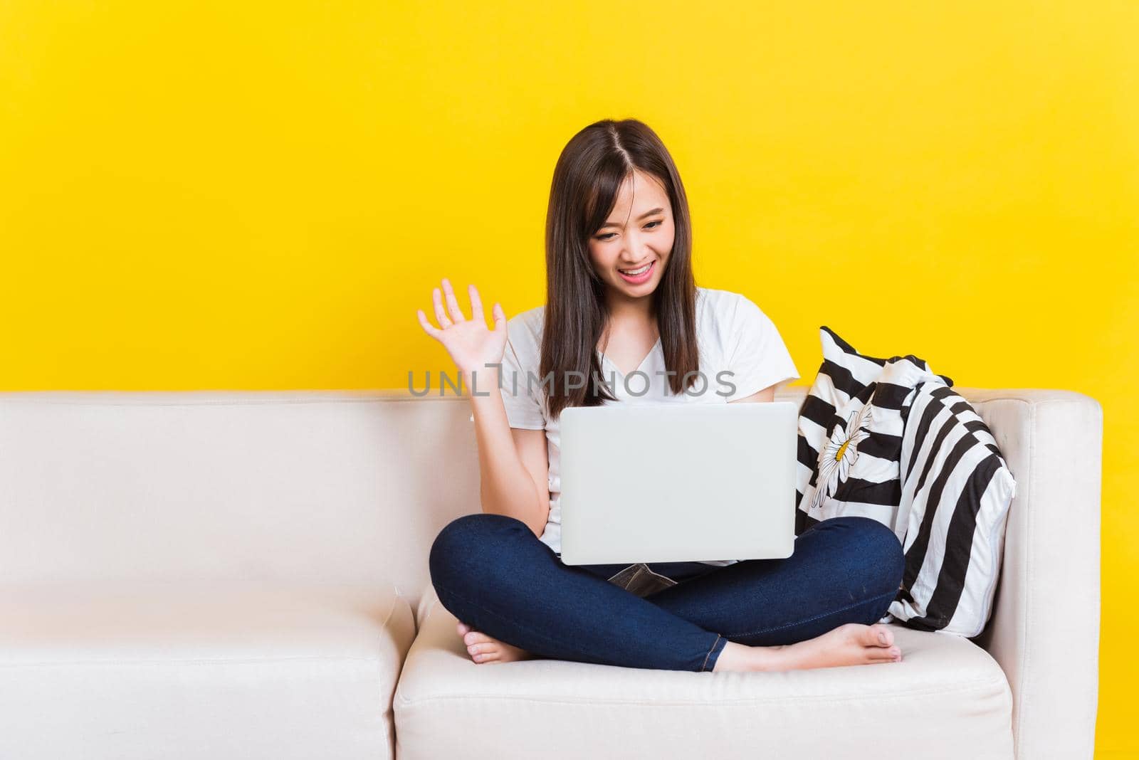 Portrait Asian of happy beautiful young woman work from home she sitting on sofa raise hand and say hello on laptop computer in house living room studio shot isolated on yellow background