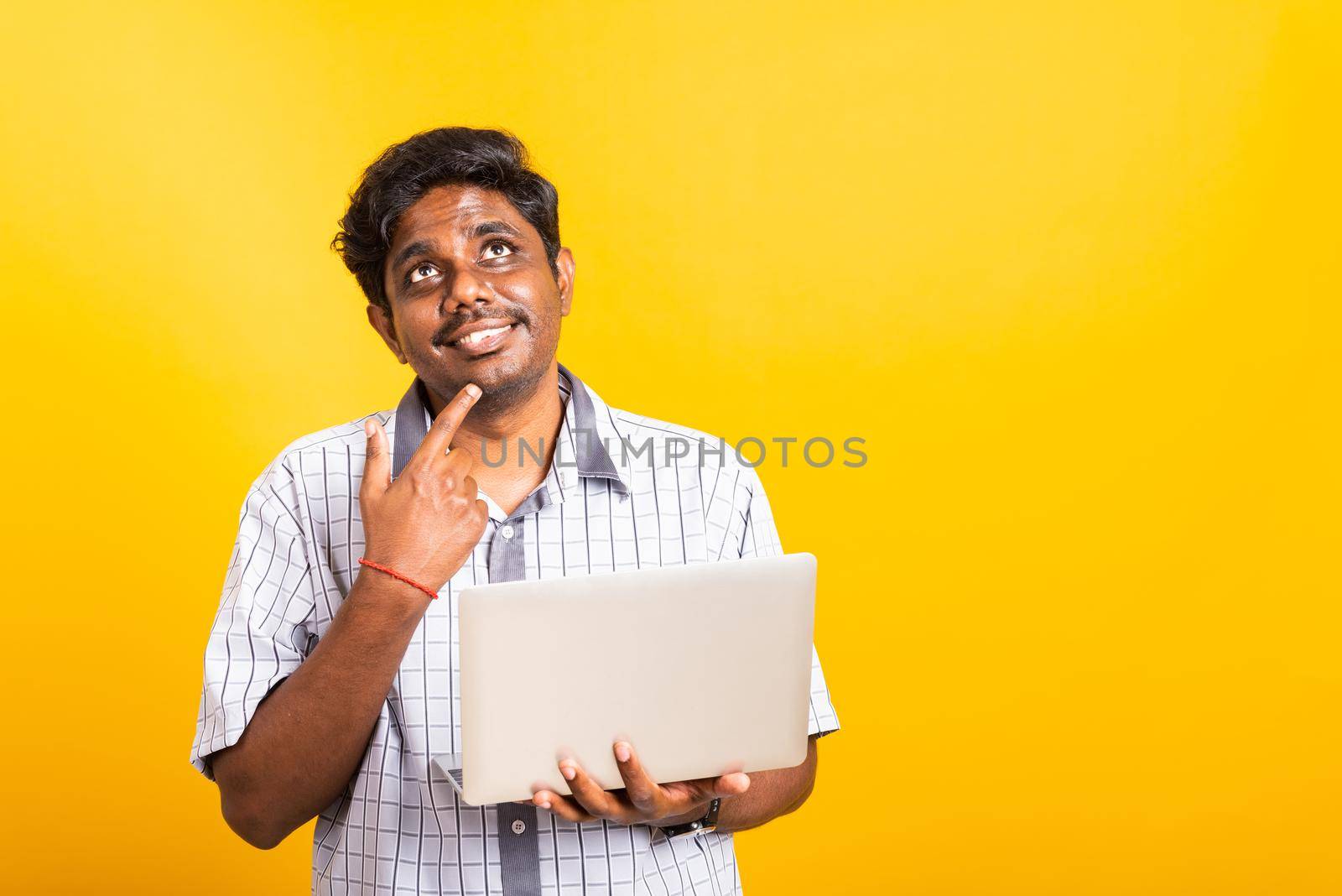 Closeup Asian happy portrait young black man holds a laptop computer hand finger handle on the chin and thinking question looking away, studio isolated on yellow background