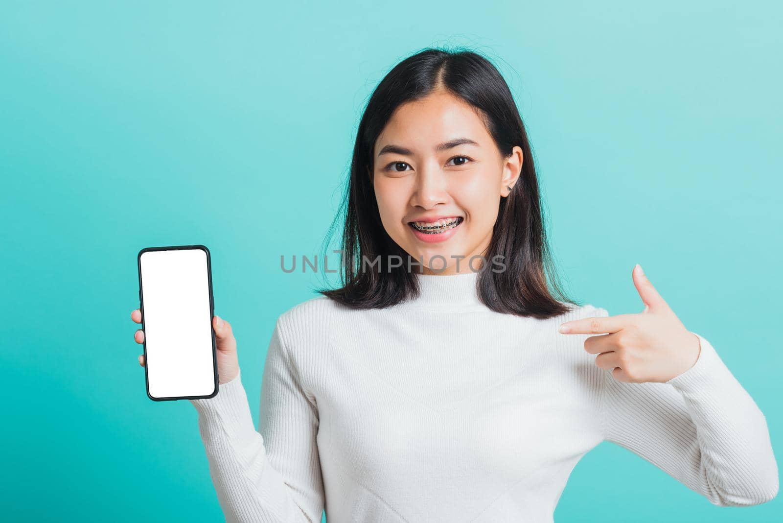 Beautiful Asian woman smile holding a smartphone on hand and pointing finger to the blank screen, female excited cheerful her show mobile phone isolated on a blue background, Technology concept