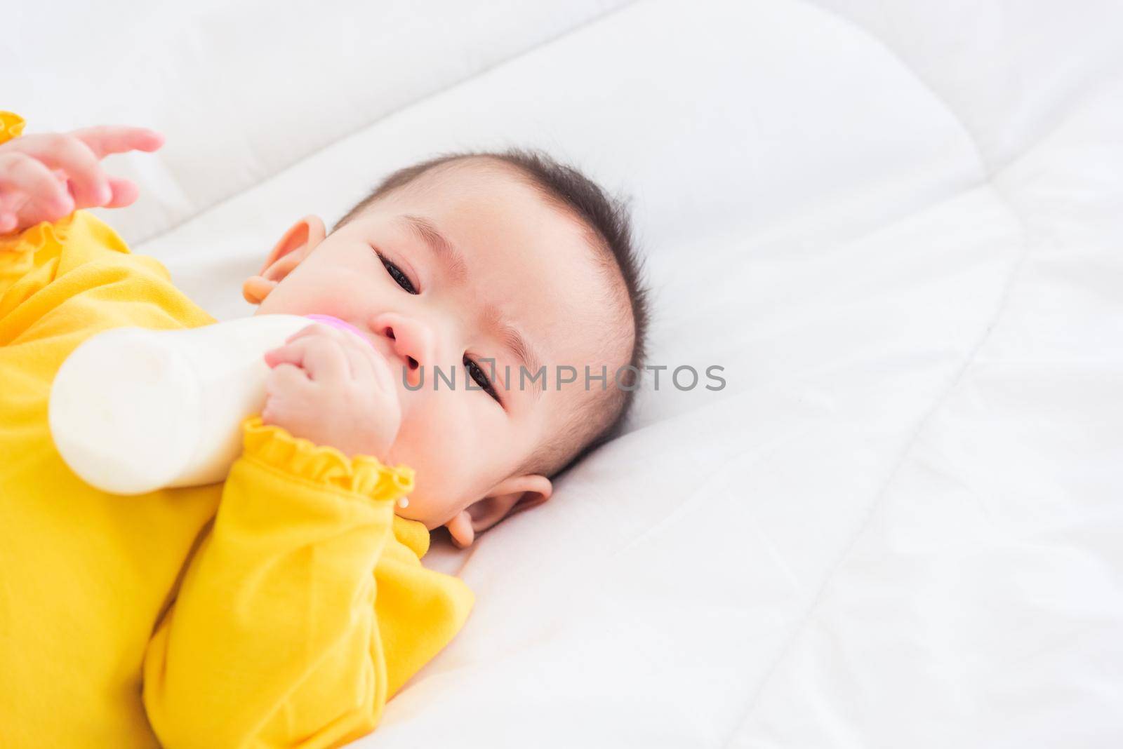 Asian beautiful little baby girl wearing a yellow dress eat milk sleeping feeding lying on the white bed, infant holding a bottle of milk, baby food concept