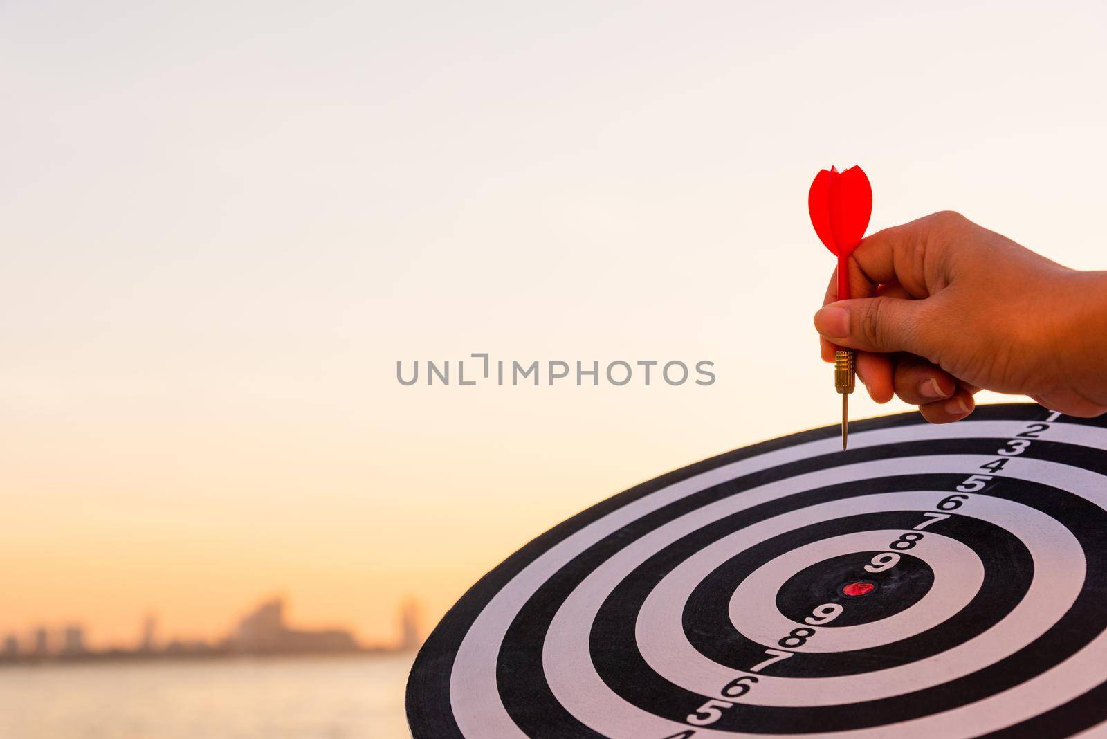 Hand hold of dart arrow on bullseye (bull's-eye) dartboard is the target of purpose challenge business at sunset, expert marketing strategy target, objective financial and goal success