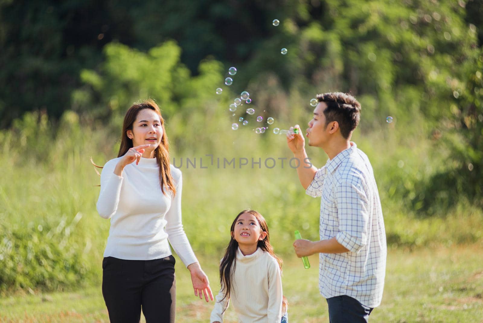 Asian family mother, father and little girl having fun together play blowing soap bubbles in park by Sorapop