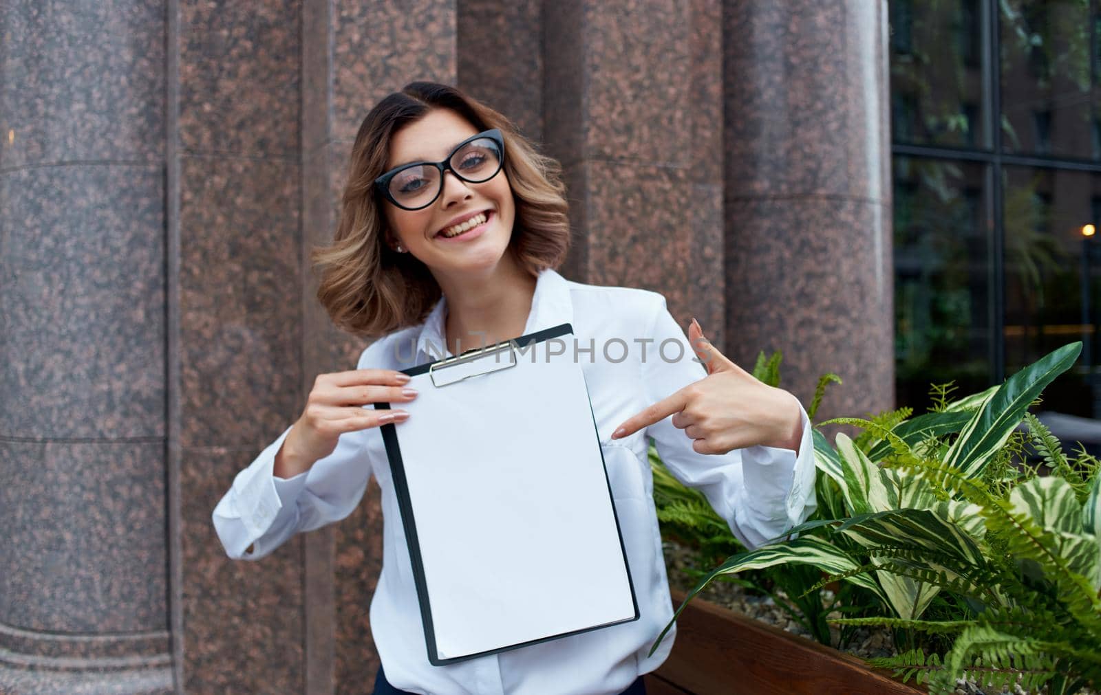 Happy business woman with documents near the building on the street in glasses and in a white shirt by SHOTPRIME