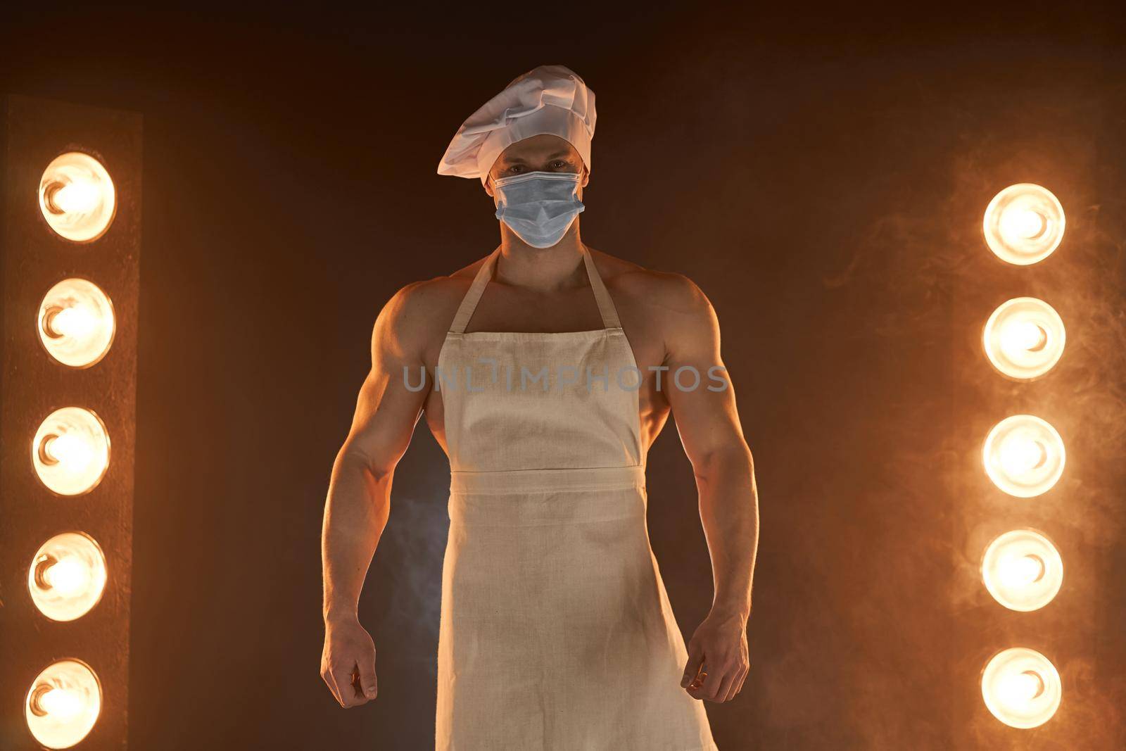 New normal concept. Portrait of muscular chef wearing protective medical mask white apron and chef hat, standing on smoky background and lamp light male housewife. husband in kitchen. brutal butcher.