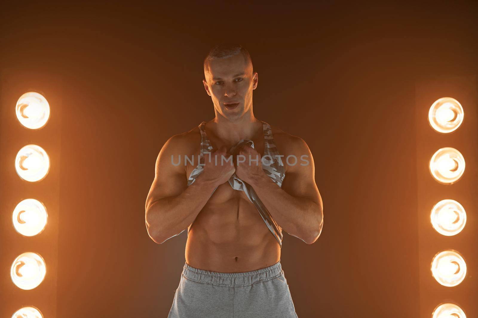 Active lifestyle concept. Professional bodybuilder picks up a t-shirt showing abs muscle and looking at camera, lamps illumination and smoke on background Sport and fitness concept