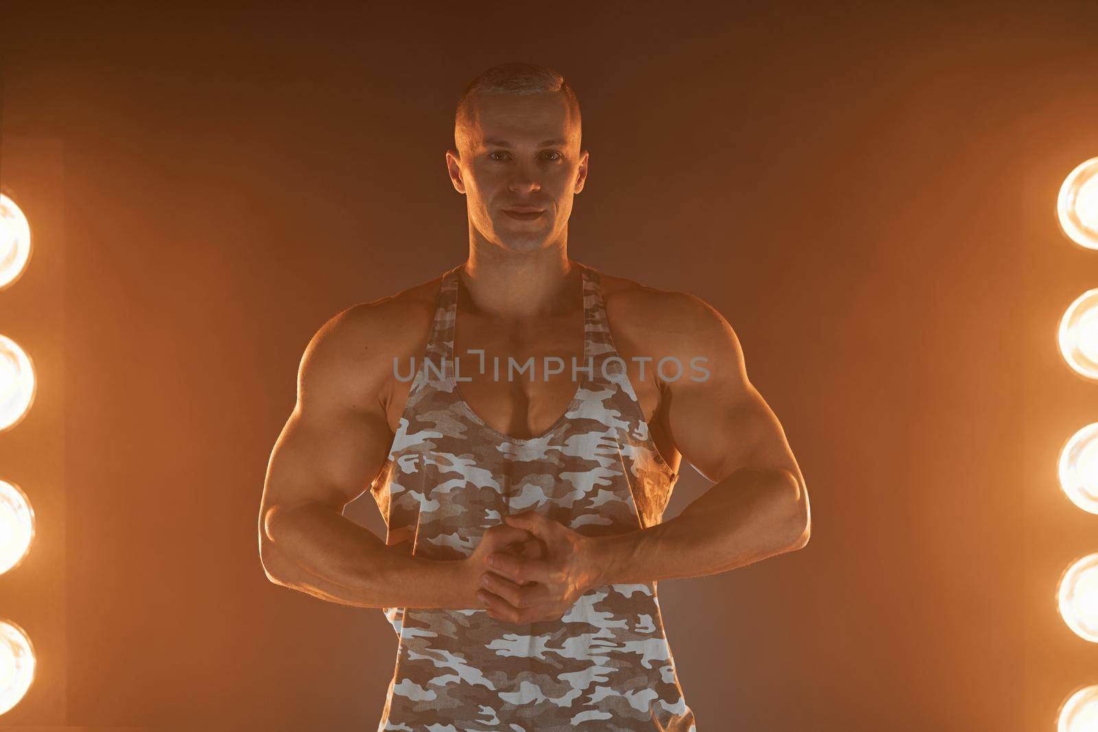 Active lifestyle concept. Professional bodybuilder showing biceps and smiling at camera, lamps illumination and smoke on background