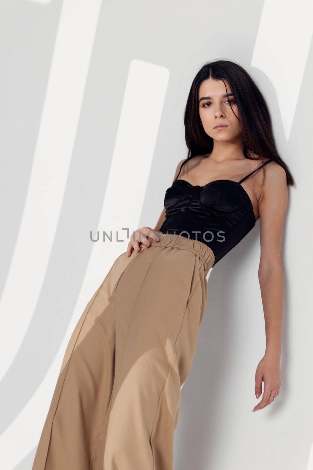 a pretty brunette in a short top in beige trousers leaned against the light wall of the room by SHOTPRIME