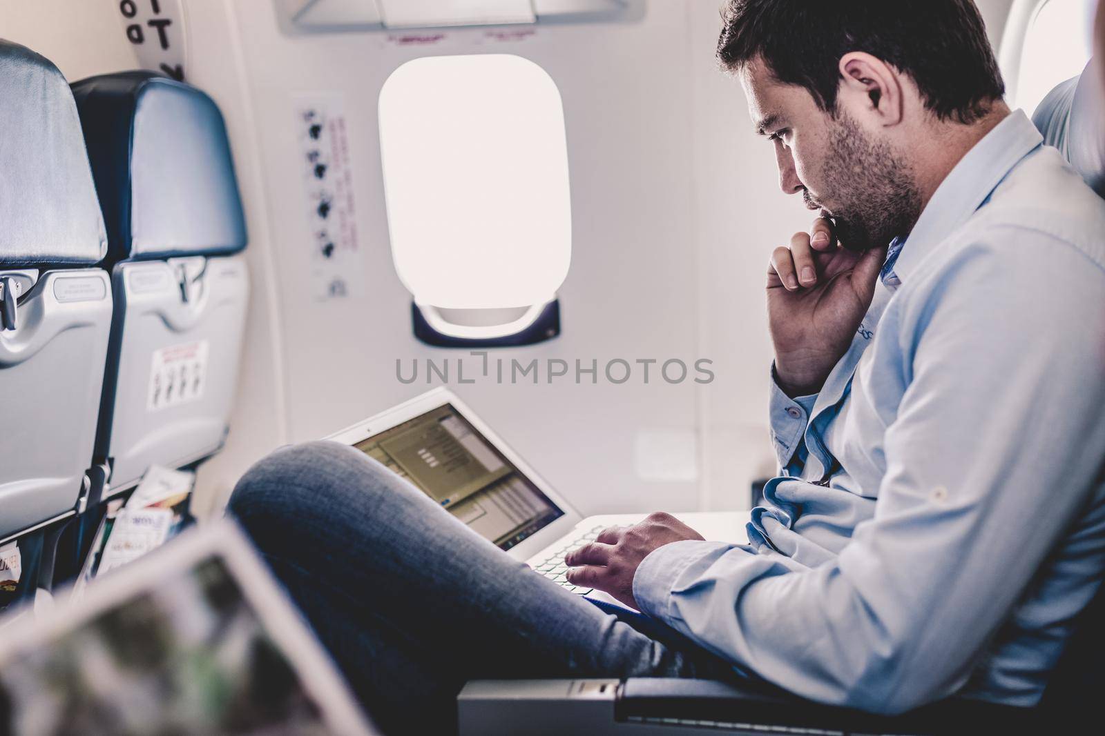 Casually dressed middle aged man working on laptop in aircraft cabin during his business travel. Shallow depth of field photo with focus on businessman eye.