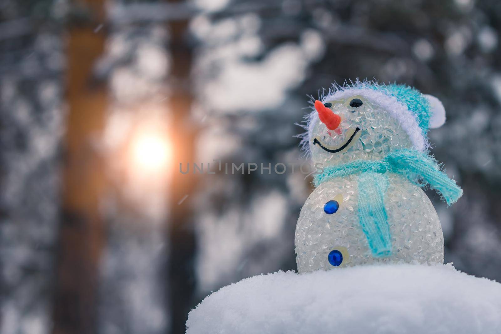 A snowman doll stands on a snowdrift against the background of a forest through which the sun breaks