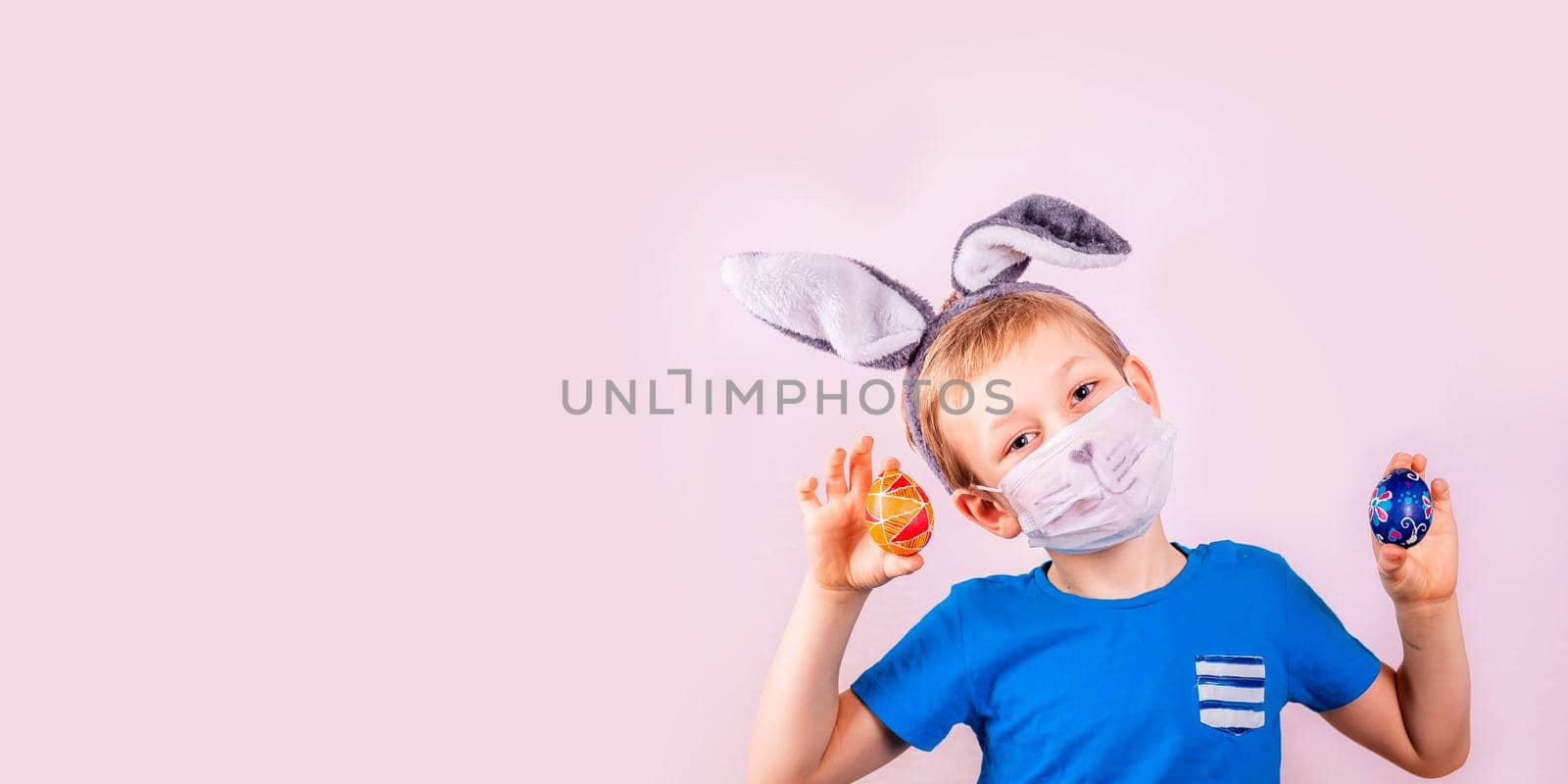 Cute little boy in rabbit bunny ears on head and protective mask with colored eggs on pink background. Cheerful smiling happy child. Covid Easter holiday banner
