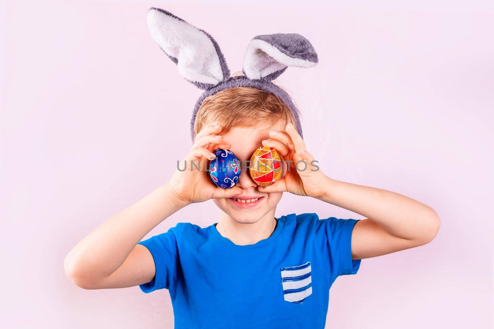 Cute little boy in rabbit bunny ears on head closing his eyes with colored eggs on pink background. Cheerful smiling happy child. Easter holiday
