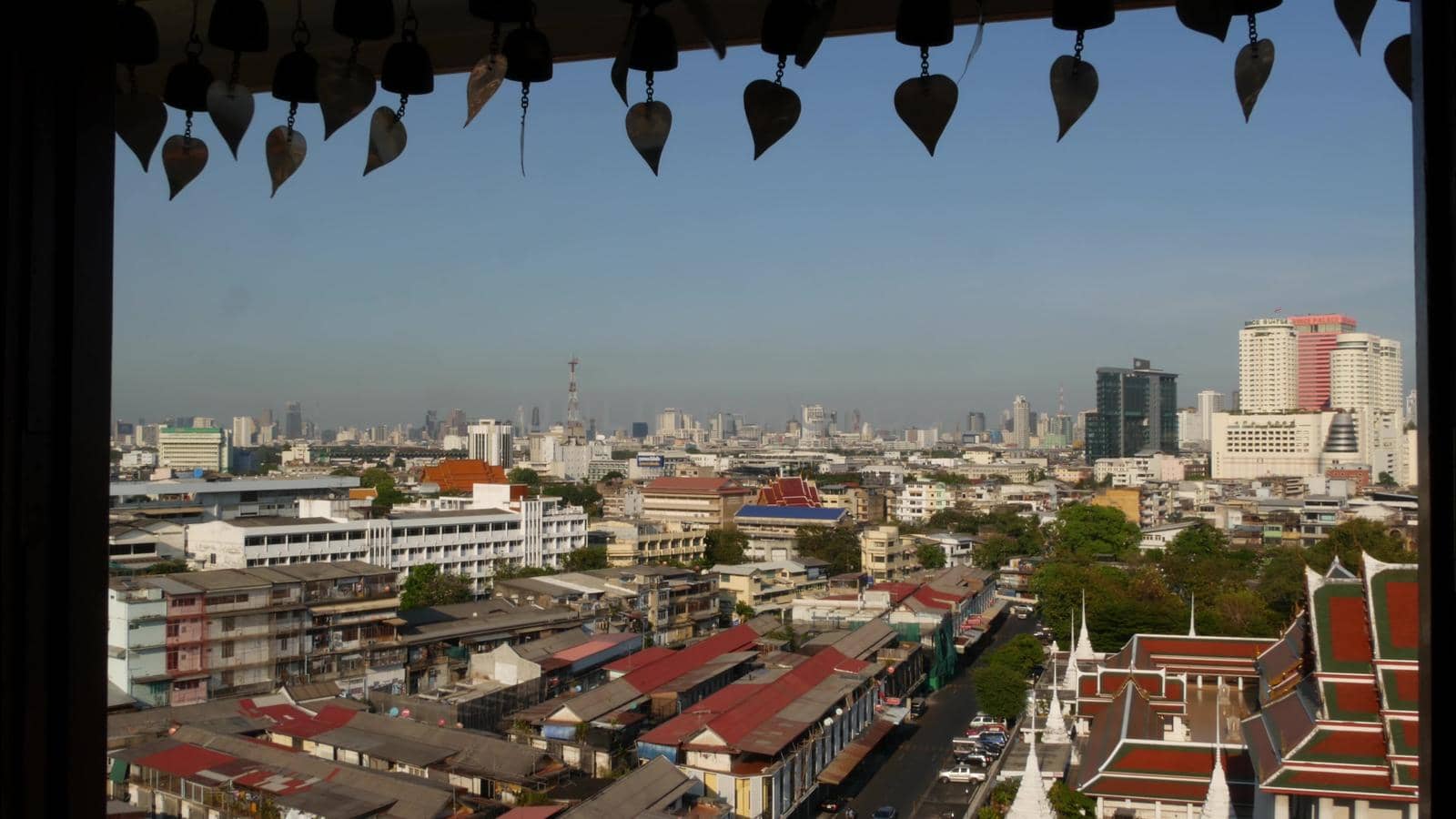 Cityscape of modern oriental town. View from the window of houses roofs on streets of majestic Bangkok from Golden Mount Temple during sunset time. Traditional metal bells with wishes hanging by DogoraSun