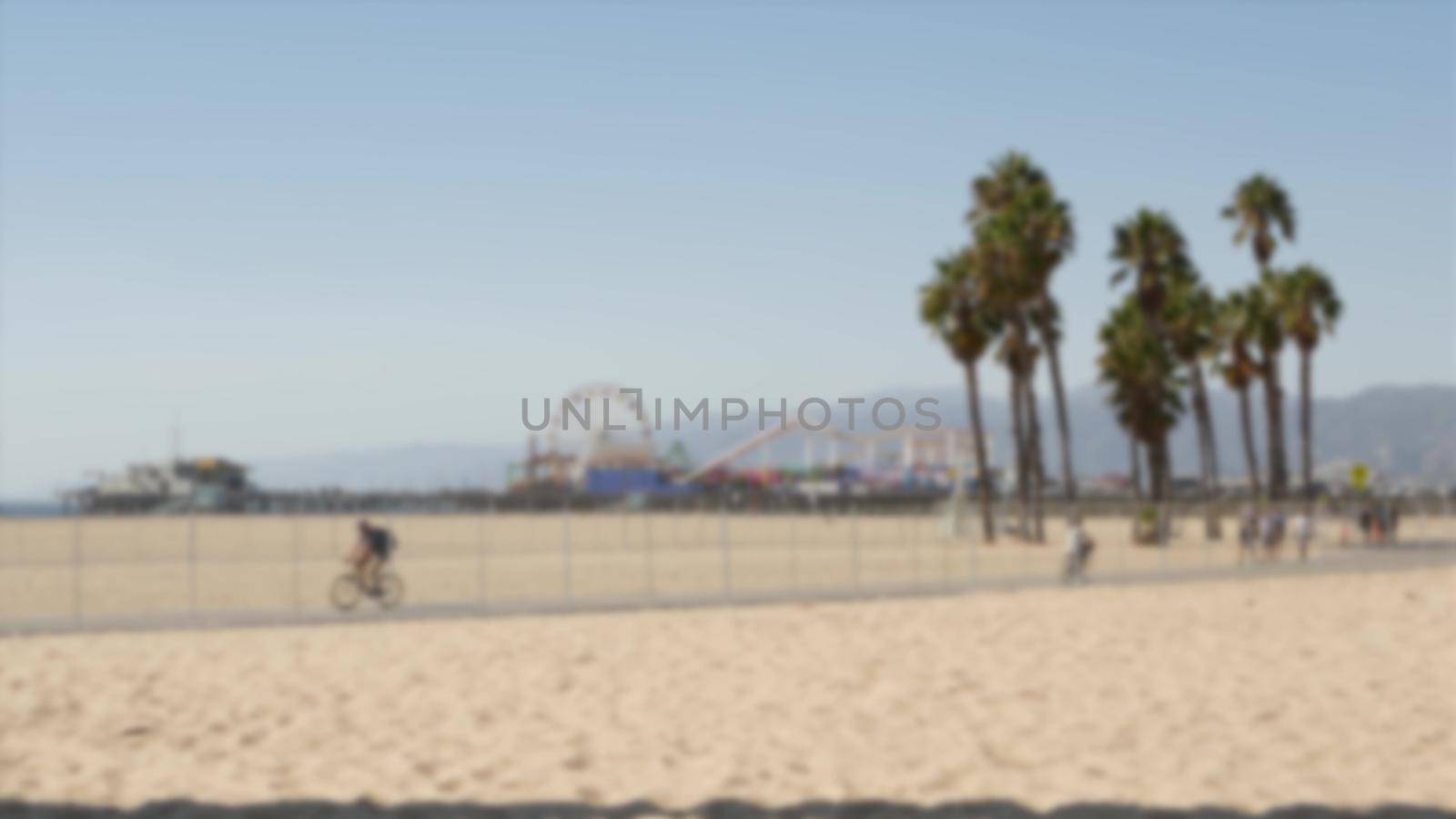 California beach aesthetic, people ride cycles on a bicycle path. Blurred, defocused background. Amusement park on pier and palms in Santa Monica american pacific ocean resort, Los Angeles CA USA.