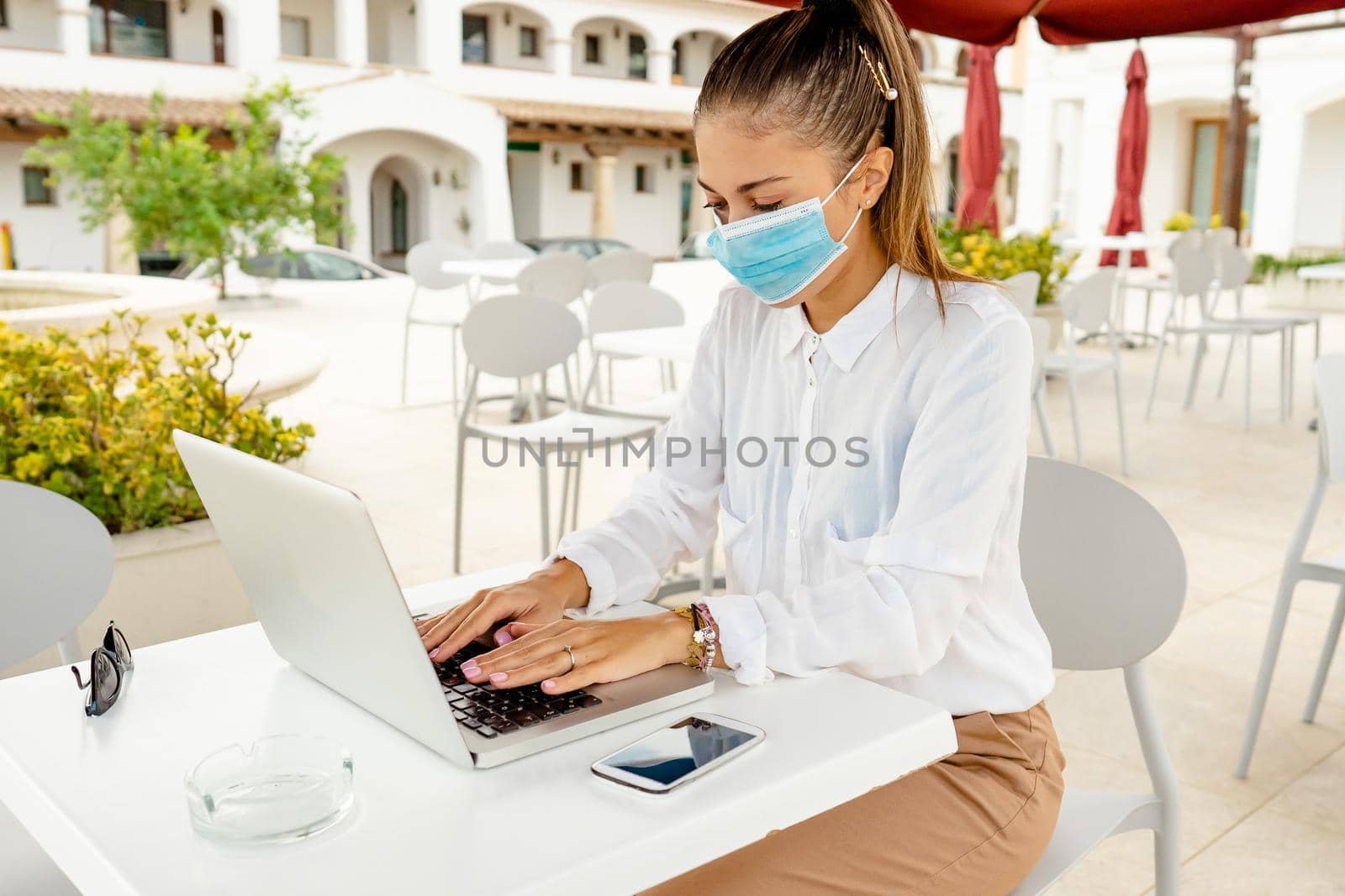 Business young woman outdoor with laptop on bar table wearing protective mask against Coronavirus pandemic. New normal freelance job activity self entrepreneur working everywhere with mobile internet