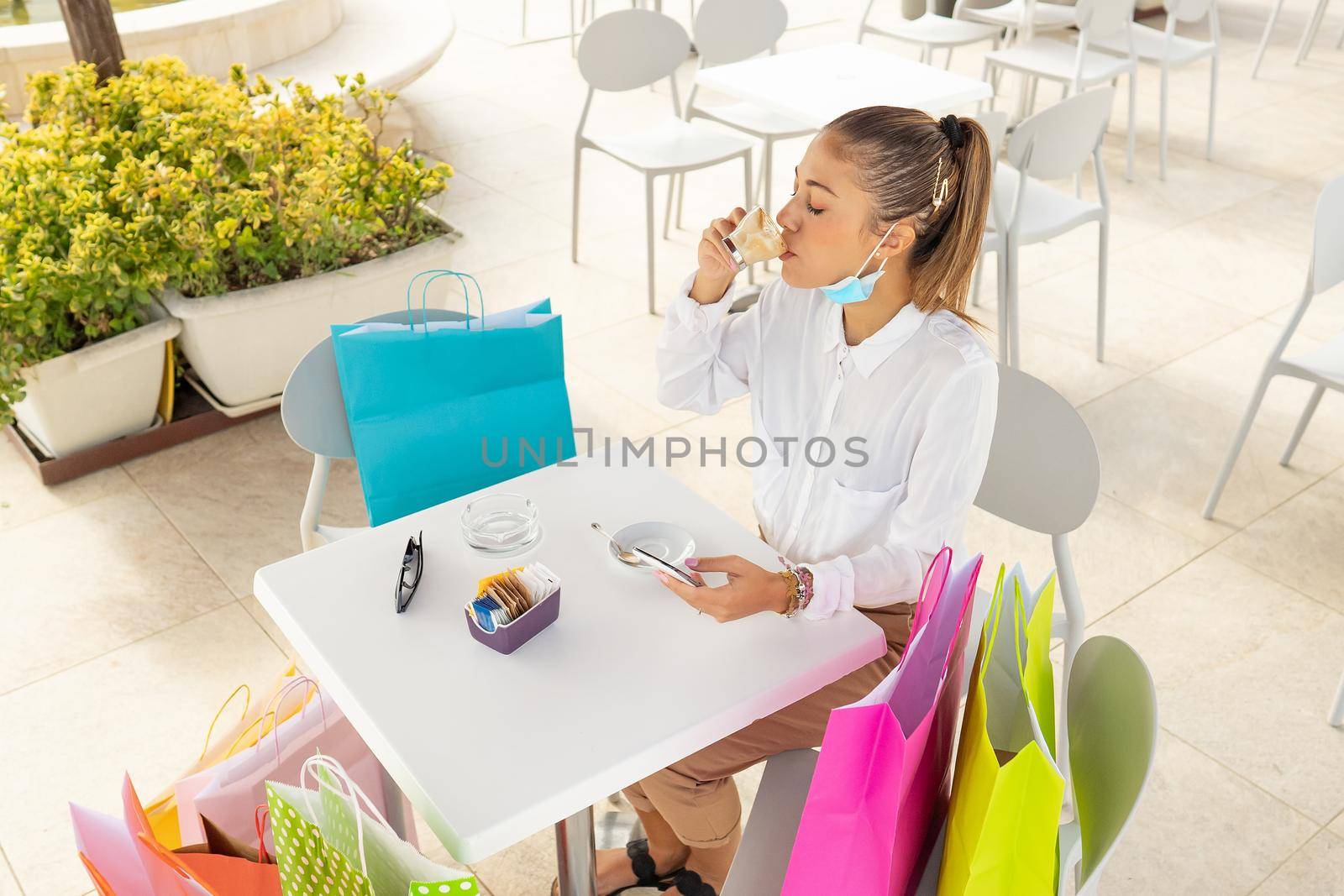 Modern young stylish woman drinking coffe outdoor at bar table with colored shopping bags using smartphone checking online best price. New normal alone female shopaholic at cafe restaurant with mask by robbyfontanesi