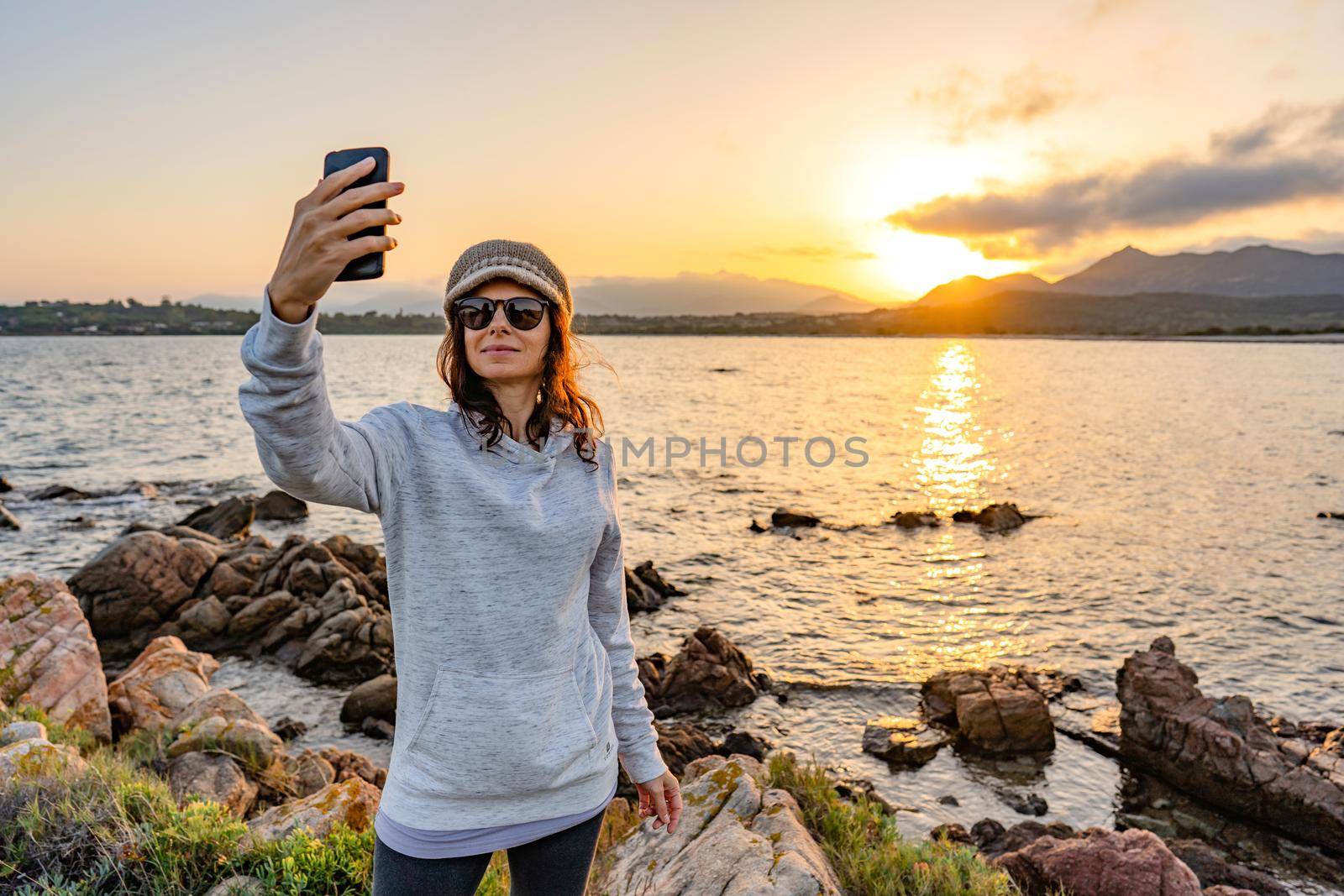 Young Caucasian woman taking a self-portrait looking at camera with sunglasses and wool hat on ocean sea resort at sunset or dawn. Solo female traveller having fun sharing her photo on social network