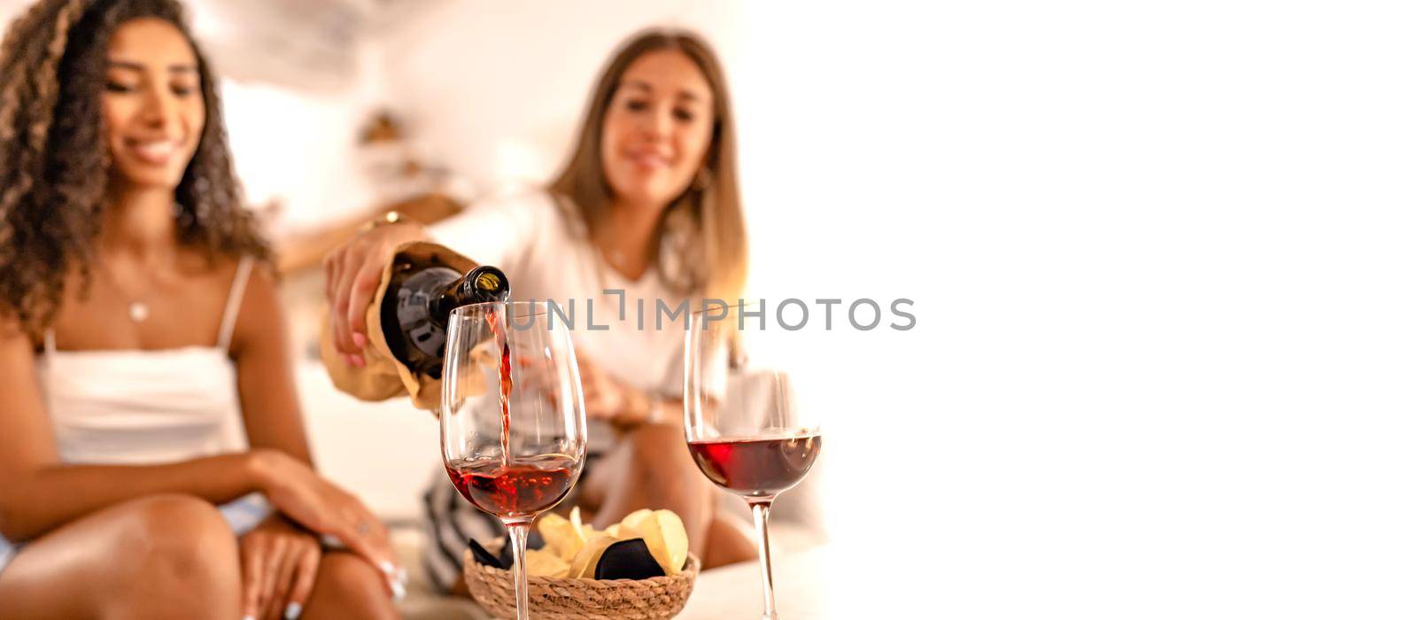 Two happy female friends celebrating at home pouring red wine in the glasses in selective focus effect and copy space. Young Caucasian woman toasting with her best Hispanic friend drinking alcohol by robbyfontanesi