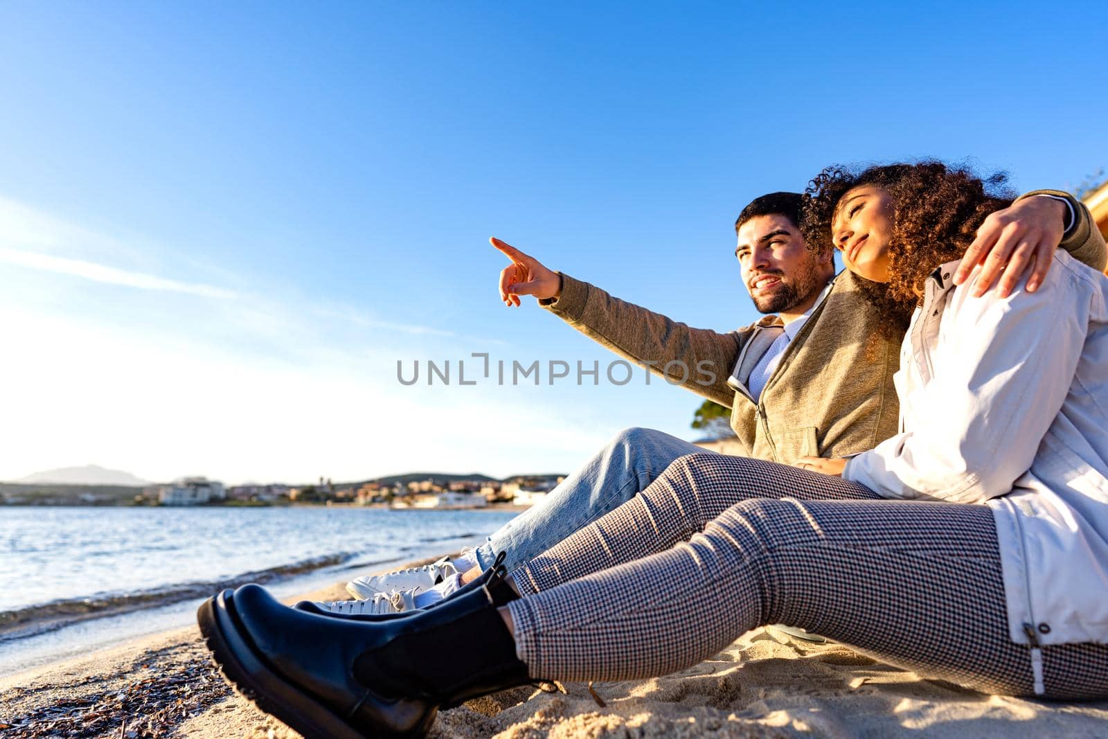 Affectionate young couple in love sitting on seashore near the ocean water next to each others looking at the sunset. Handsome man embracing girlfriend pointing to horizon in winter vacation travel