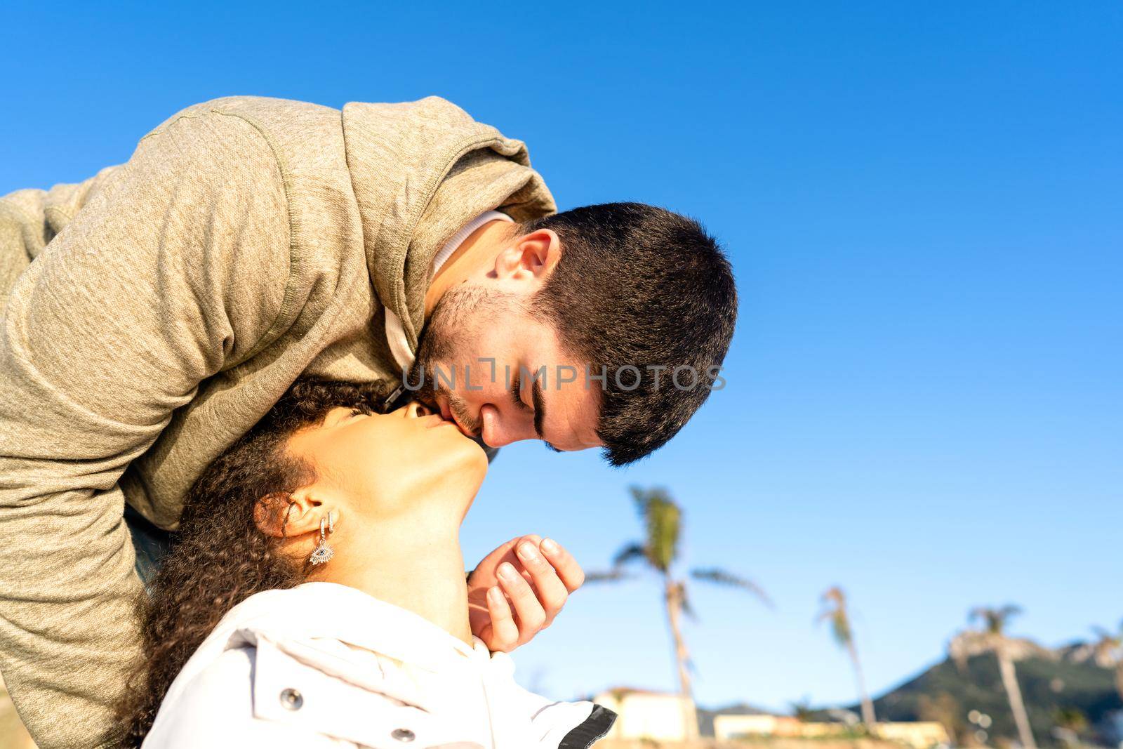 Handsome guy kissing her african-american black girlfriend approaching her mouth from above. Mixed race young couple in love romance vacation scene in sea or ocean resort with copy space in blue sky by robbyfontanesi