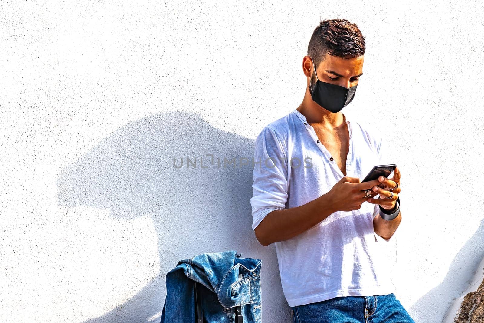 Young solo traveller leaning at a white wall with trolley bag using smartphone chatting online waiting bus or train wearing Coronavirus black protective mask. Modern guy in bright color vivid effect by robbyfontanesi