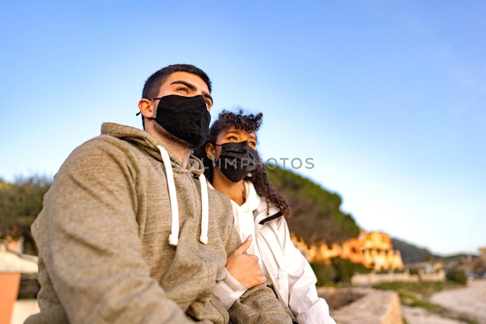MIxed-race afro-american girl embracing her Caucasian boyfriend sitting outdoor in sea resort looking the sunset wearing black protective mask against Coronavirus pandemic. New normal vacation travel